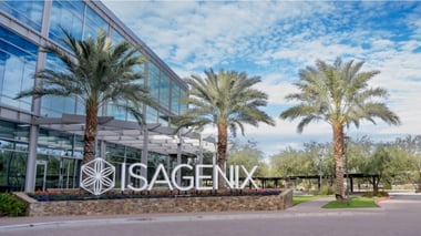 Modern corporate office building with a glass exterior and palm trees, featuring an ISAGENIX logo sign at the entrance and landscaped surroundings.
