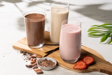 Three glasses of blended shakes in chocolate, vanilla, and strawberry flavors arranged on a wooden board with fruit and cocoa ingredients.
