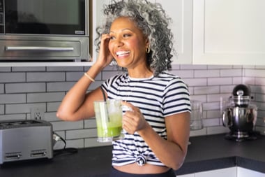 Person standing in a kitchen holding a glass mug filled with a green blended drink, with kitchen appliances visible in the background.