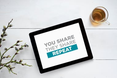 Tablet displayed on a white table showing the text ‘YOU SHARE THEY SHARE REPEAT,’ with a glass of tea and plant stems nearby.