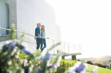 Two people walking along an outdoor walkway with a railing, overlooking a landscaped hillside and modern building in bright daylight.