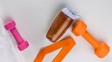 Isagenix shaker bottle filled with a brown blended drink surrounded by orange and pink dumbbells and resistance bands on a white surface.