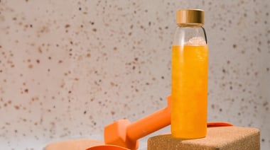 Clear bottle filled with a yellow beverage placed on a cork block beside an orange dumbbell on a textured surface.