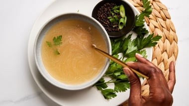 Top-down view of a bowl of light broth garnished with herbs, with a hand holding a spoon and small bowls of pepper and green onions nearby.