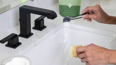 Hand pouring pure sparkle bathroom cleaner into a sink while scrubbing with a brush.