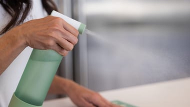 Woman spraying solution onto countertop with an Isagenix green, glass spray bottle. 