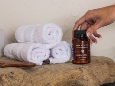 Hand placing a Hair Revival supplement bottle next to rolled white towels on a wooden surface.