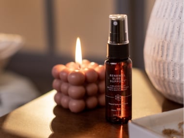 Bottle of Renewal Sleep Support melatonin mist on a bedside table beside a lit candle.