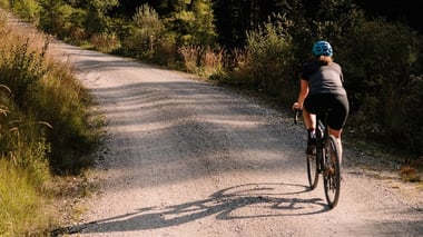Person cycling on a sunlit gravel road surrounded by trees.