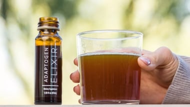 Bottle of Adaptogen Elixir beside a glass of amber-colored beverage.