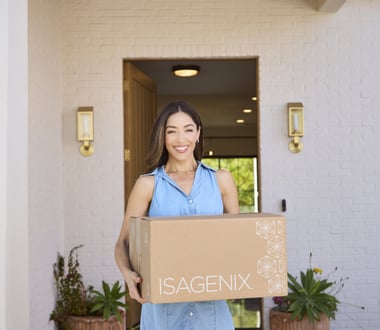 Person standing at a front doorway holding a large cardboard box labeled ‘ISAGENIX,’ with a home exterior and plants visible.