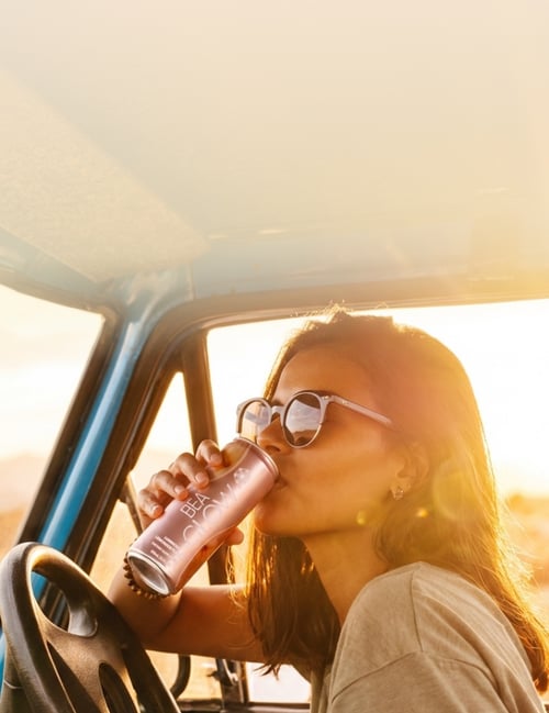 Woman drinking a can of BEA Glow in the front seat of her car with a desert sunset in the background.
